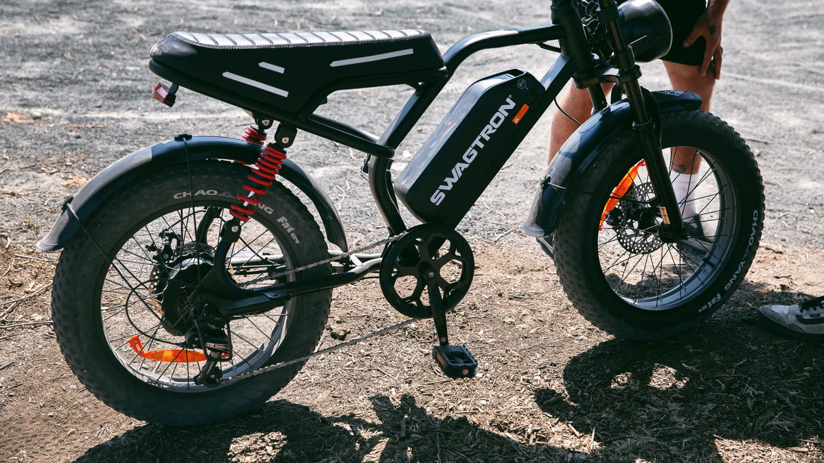 Side view of a SWAGTRON UrbanCruise e-bike parked on a sunny dirt lot, showing its bench seat, red rear shock and fat tires