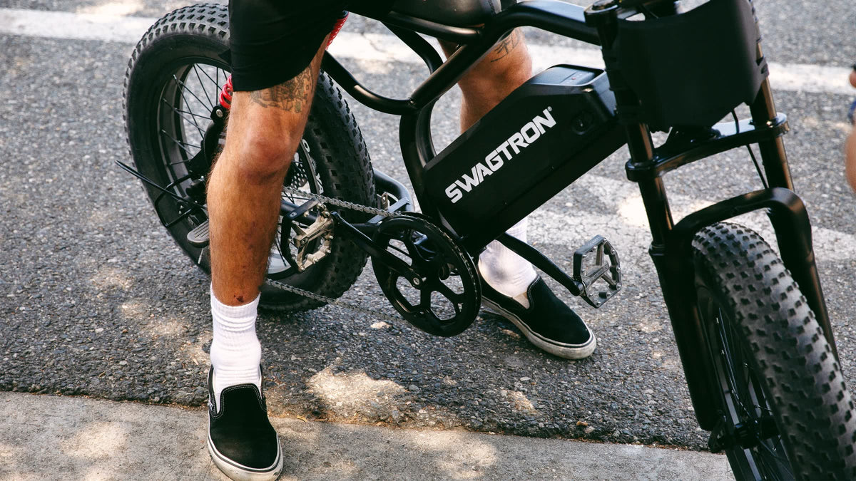 Rider in shorts and white socks straddling a SWAGTRON UrbanCruise e-bike on a sunny street, with the SWAGTRON wordmark on its battery