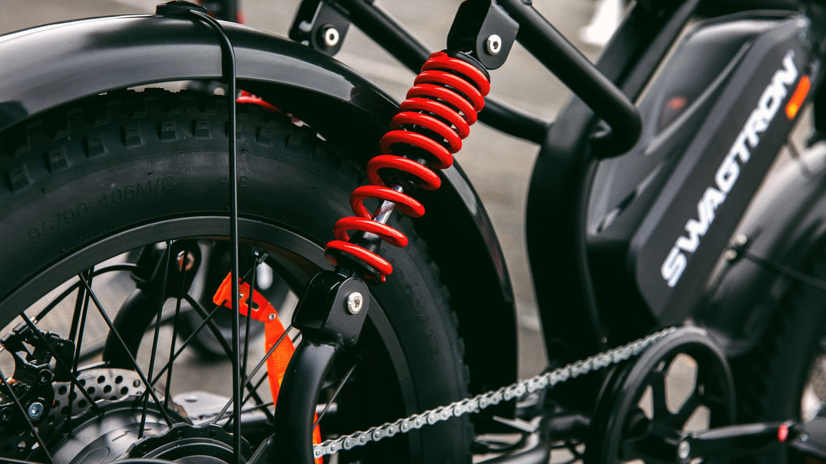 Close-up of the SWAGTRON UrbanCruise e-bike's red coil-over rear shock, fat rear tire and battery with the SWAGTRON wordmark