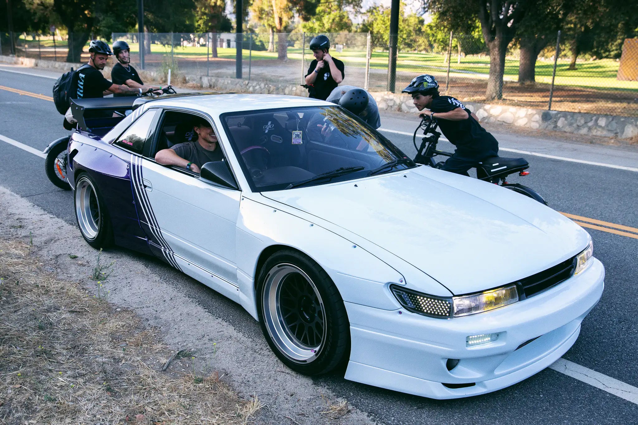 The white-and-purple Nissan S13 drift car on a park road, riders on e-bikes beside it.